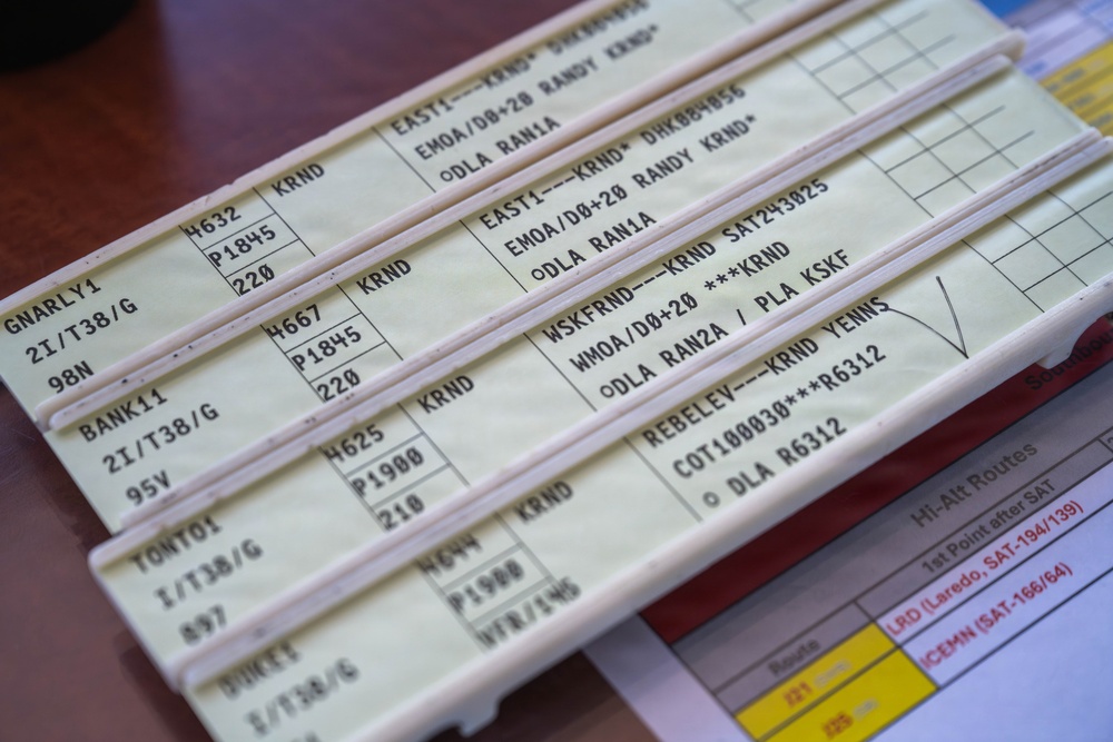 The image shows a set of aviation flight progress strips used in air traffic control, arranged in a strip holder on a desk. Each strip contains coded flight information including aircraft identifiers, altitudes, routes, and waypoints. A reference document for Hi-Alt Routes is partially visible beneath the strips.
