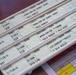 The image shows a set of aviation flight progress strips used in air traffic control, arranged in a strip holder on a desk. Each strip contains coded flight information including aircraft identifiers, altitudes, routes, and waypoints. A reference document for Hi-Alt Routes is partially visible beneath the strips.