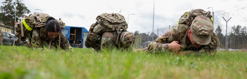 Three military soldiers in full combat gear and rucksacks are performing a low crawl exercise on a grass field at what appears to be a military installation. They are wearing multicam/OCP uniforms and carrying large tactical backpacks while crawling on their hands and knees. The setting appears to be a training event or physical fitness assessment at a base facility.