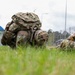 Three military soldiers in full combat gear and rucksacks are performing a low crawl exercise on a grass field at what appears to be a military installation. They are wearing multicam/OCP uniforms and carrying large tactical backpacks while crawling on their hands and knees. The setting appears to be a training event or physical fitness assessment at a base facility.