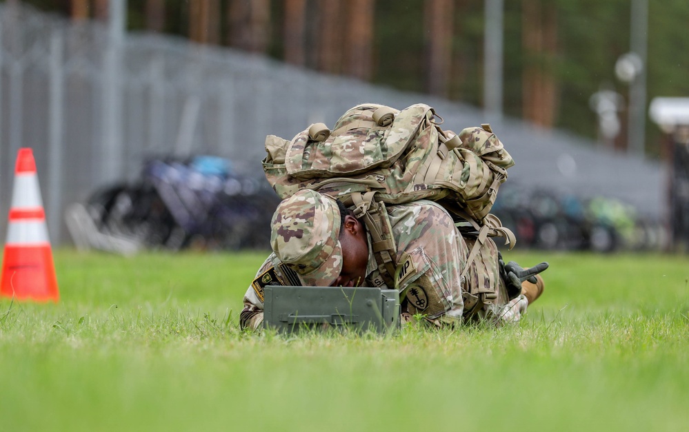 A U.S. Army soldier in full combat gear, including a large rucksack, is lying prone on a grass field closely examining or working on a military ammunition can. The soldier is wearing MultiCam (OCP) camouflage uniform with visible unit patches and appears to be engaged in a training exercise or competition event. An orange traffic cone is visible in the background, suggesting a marked course or lane.