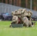 A U.S. Army soldier in full combat gear, including a large rucksack, is lying prone on a grass field closely examining or working on a military ammunition can. The soldier is wearing MultiCam (OCP) camouflage uniform with visible unit patches and appears to be engaged in a training exercise or competition event. An orange traffic cone is visible in the background, suggesting a marked course or lane.