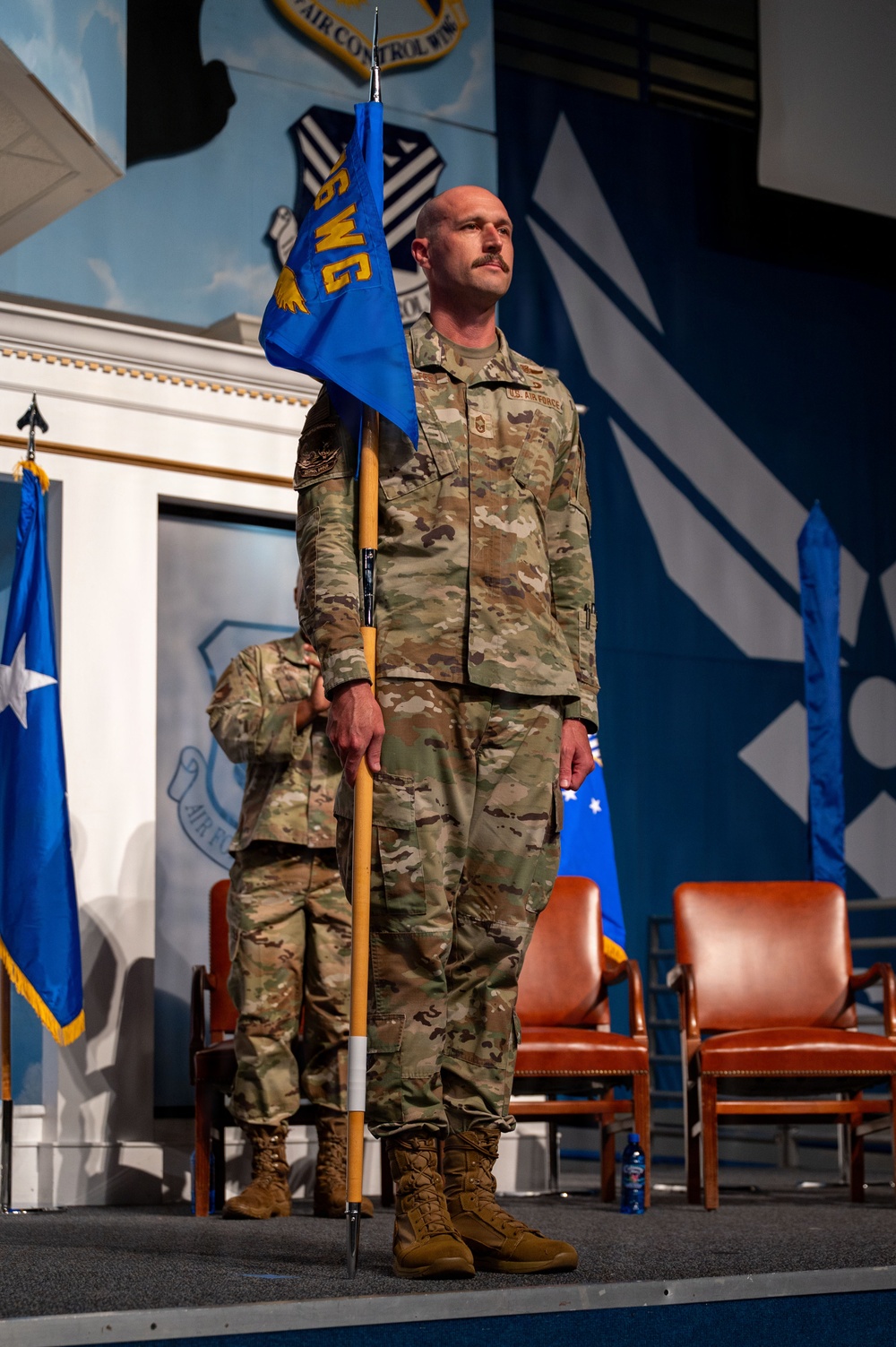 A U.S. Air Force service member in OCP (Operational Camouflage Pattern) uniform stands at attention on a stage holding a blue guidon/flag during what appears to be a formal military ceremony. The individual is bald with a mustache and displays rank insignia on their uniform. The setting features Air Force emblems, murals, and ceremonial flags in the background.