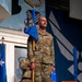 A U.S. Air Force service member in OCP (Operational Camouflage Pattern) uniform stands at attention on a stage holding a blue guidon/flag during what appears to be a formal military ceremony. The individual is bald with a mustache and displays rank insignia on their uniform. The setting features Air Force emblems, murals, and ceremonial flags in the background.
