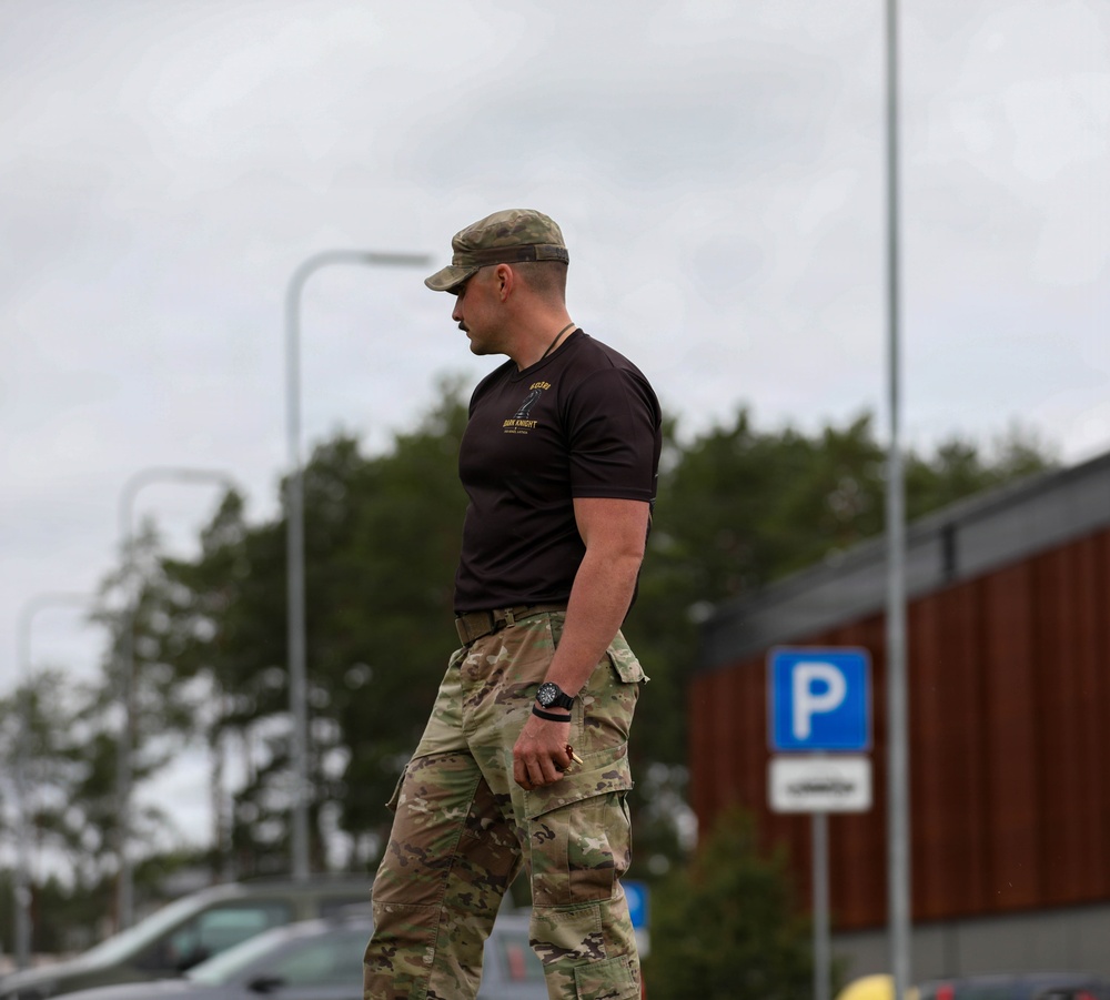 A muscular male individual wearing military-style camouflage pants, a dark t-shirt with text/insignia, and a camouflage cap is walking through what appears to be a parking area. He is wearing a tactical watch and appears to be in a relaxed but purposeful stride. The background shows trees, street lights, parked vehicles, and a blue parking sign consistent with a Northern European setting.