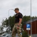 A muscular male individual wearing military-style camouflage pants, a dark t-shirt with text/insignia, and a camouflage cap is walking through what appears to be a parking area. He is wearing a tactical watch and appears to be in a relaxed but purposeful stride. The background shows trees, street lights, parked vehicles, and a blue parking sign consistent with a Northern European setting.