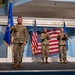 Three U.S. Air Force personnel in OCP (Operational Camouflage Pattern) uniforms are standing on a stage at the Museum of Aviation at Robins Air Force Base. One service member on the left holds a blue unit guidon, while two others in the center and right hold an unfolded American flag. The setting appears to be a formal military ceremony.
