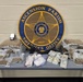 This is an official law enforcement evidence display photograph from the Ascension Parish Sheriff's Office in Louisiana. The image shows seized contraband, currency, and a firearm arranged on a table in front of the agency's official seal. This type of photograph is typically used for press releases or official documentation of drug-related arrests.