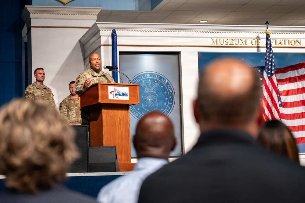 A military officer in camouflage uniform delivers a speech at a podium bearing the Museum of Aviation logo, with the 'Museum of Aviation' signage visible on the wall behind. The event appears to be a formal ceremony or official address, with an audience seated in the foreground and additional uniformed personnel standing on the stage. A U.S. flag and what appears to be a Department of the Air Force seal are visible in the background.