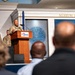A military officer in camouflage uniform delivers a speech at a podium bearing the Museum of Aviation logo, with the 'Museum of Aviation' signage visible on the wall behind. The event appears to be a formal ceremony or official address, with an audience seated in the foreground and additional uniformed personnel standing on the stage. A U.S. flag and what appears to be a Department of the Air Force seal are visible in the background.