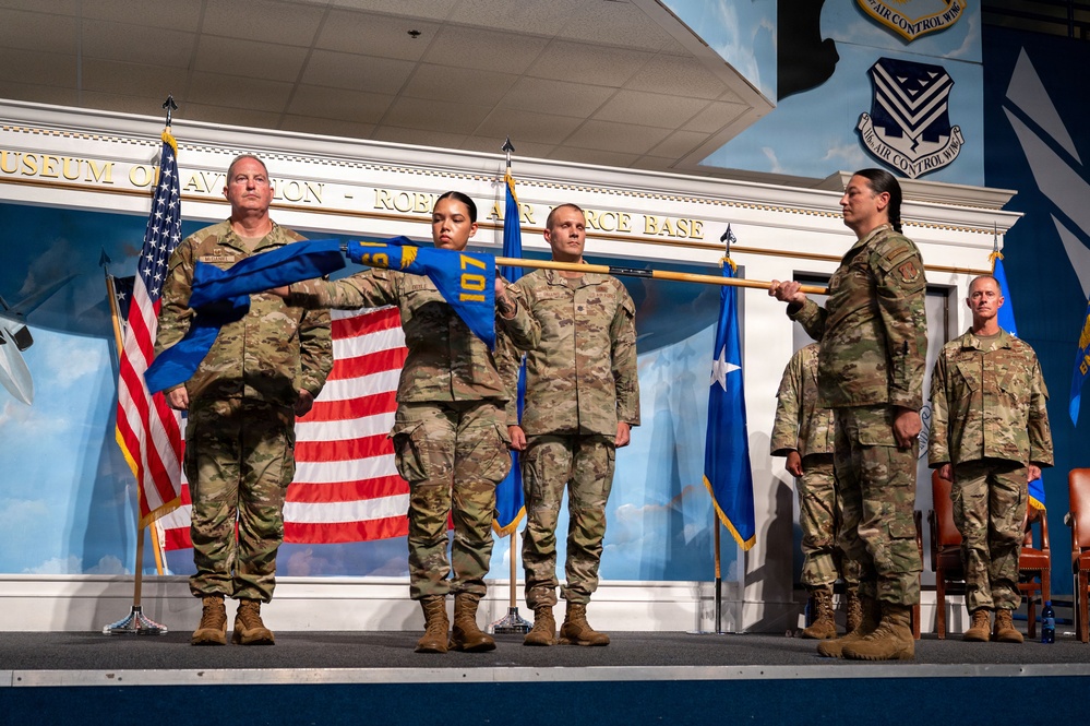 A military ceremony is taking place on a stage at what appears to be the Museum of Aviation at Robins Air Force Base. Several service members in OCP (Operational Camouflage Pattern) uniforms are participating in what appears to be a guidon or unit colors unfurling/casing ceremony. A blue unit guidon with the number '107' visible is being displayed or transferred between personnel.