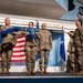 A military ceremony is taking place on a stage at what appears to be the Museum of Aviation at Robins Air Force Base. Several service members in OCP (Operational Camouflage Pattern) uniforms are participating in what appears to be a guidon or unit colors unfurling/casing ceremony. A blue unit guidon with the number '107' visible is being displayed or transferred between personnel.