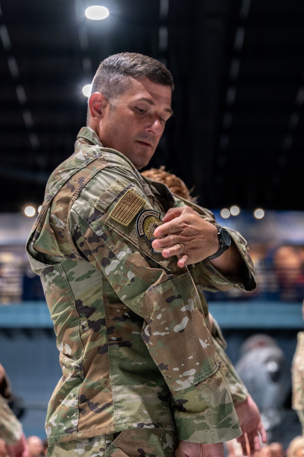 A male military service member wearing OCP (Operational Camouflage Pattern) uniform is engaged in what appears to be a reunion embrace, holding a child. He is wearing a US flag patch and a unit patch on his left sleeve, along with a dark wristwatch. The setting appears to be an indoor venue such as a hangar or large event hall.