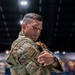 A male military service member wearing OCP (Operational Camouflage Pattern) uniform is engaged in what appears to be a reunion embrace, holding a child. He is wearing a US flag patch and a unit patch on his left sleeve, along with a dark wristwatch. The setting appears to be an indoor venue such as a hangar or large event hall.