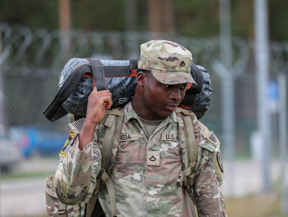 A U.S. Army soldier in Operational Camouflage Pattern (OCP) uniform is carrying a heavy load on their shoulder, appearing to be engaged in a physical training or ruck march exercise. The soldier's name tape reads 'VEDA' and rank insignia indicates Private First Class. The background shows a military installation with concertina wire fencing and tree line.