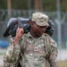 A U.S. Army soldier in Operational Camouflage Pattern (OCP) uniform is carrying a heavy load on their shoulder, appearing to be engaged in a physical training or ruck march exercise. The soldier's name tape reads 'VEDA' and rank insignia indicates Private First Class. The background shows a military installation with concertina wire fencing and tree line.