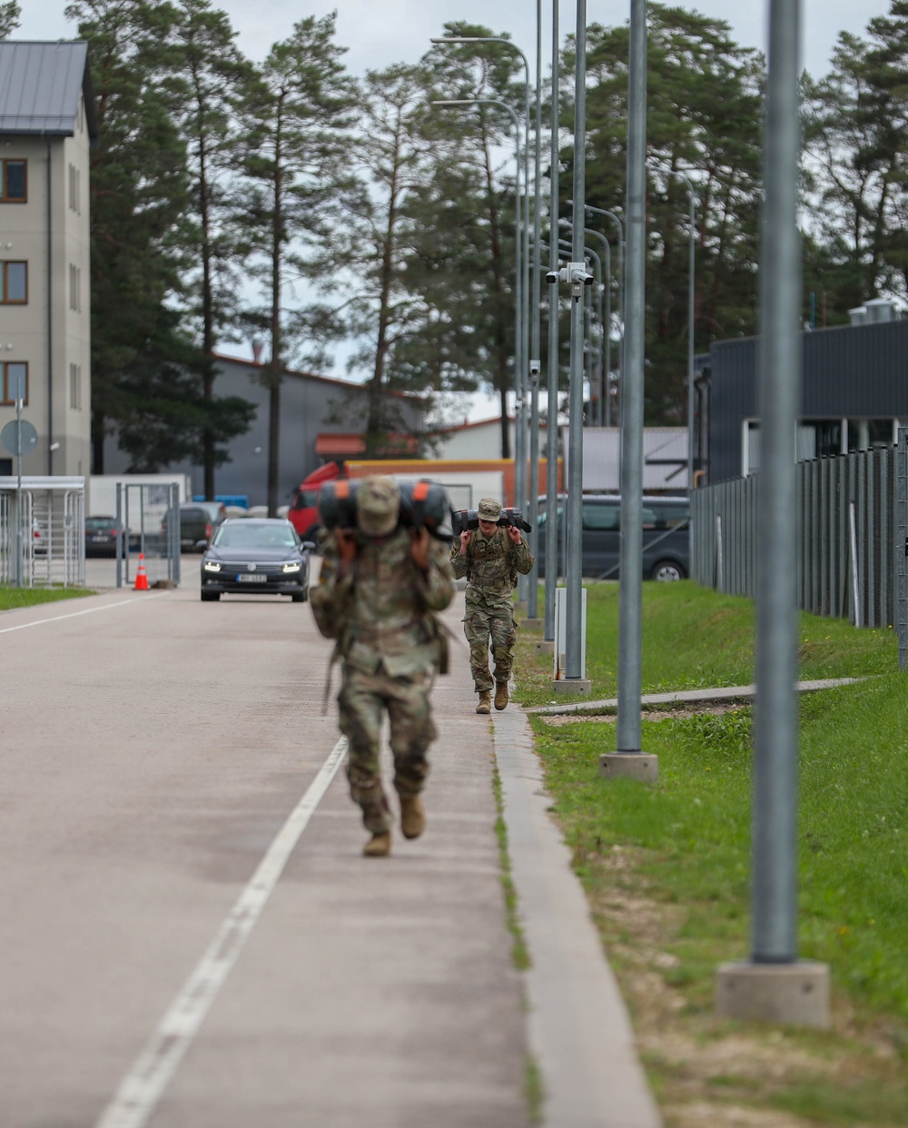 Two soldiers in camouflage uniforms (OCP/MultiCam pattern) are conducting a ruck march along a paved road on what appears to be a military installation. Both carry large rucksacks and are moving in the same direction away from the camera. The setting includes modern military base infrastructure with street lamps, fencing, buildings, and parked vehicles.