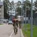 Two soldiers in camouflage uniforms (OCP/MultiCam pattern) are conducting a ruck march along a paved road on what appears to be a military installation. Both carry large rucksacks and are moving in the same direction away from the camera. The setting includes modern military base infrastructure with street lamps, fencing, buildings, and parked vehicles.