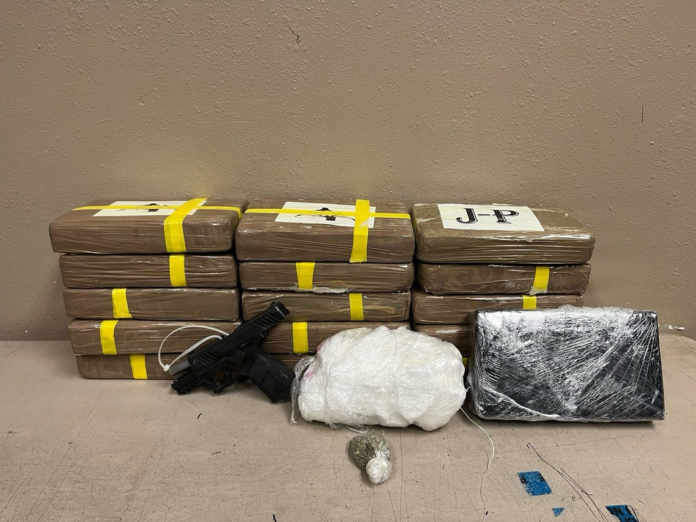 The image depicts what appears to be a law enforcement evidence display showing seized contraband items arranged on a surface against a beige wall. Multiple brown-wrapped rectangular packages bound with yellow tape are stacked in two rows, with labels visible including 'J-P'. In front of the packages are additional items including a handgun, a white mesh bag containing a substance, a small green organic material, and a dark-wrapped package.