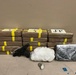 The image depicts what appears to be a law enforcement evidence display showing seized contraband items arranged on a surface against a beige wall. Multiple brown-wrapped rectangular packages bound with yellow tape are stacked in two rows, with labels visible including 'J-P'. In front of the packages are additional items including a handgun, a white mesh bag containing a substance, a small green organic material, and a dark-wrapped package.
