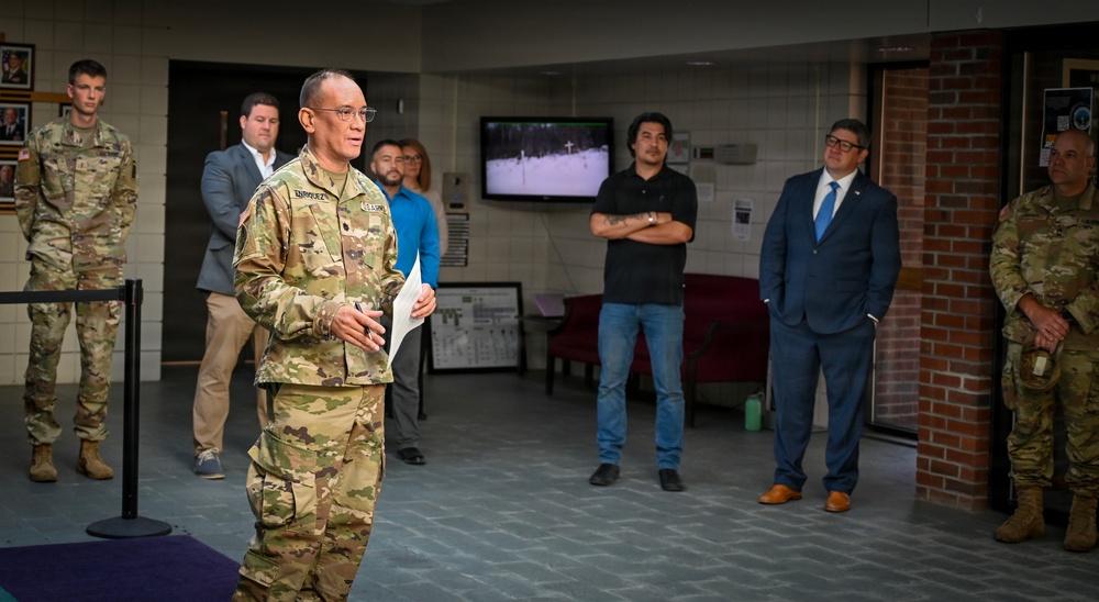 A military officer in Army Combat Uniform (ACU/OCP pattern) appears to be addressing a small group in what looks like a military building lobby or reception area. The speaker is holding papers and appears to be giving a briefing or presentation to a mixed audience of military personnel and civilians. The setting suggests an official military event or visit.