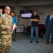 A military officer in Army Combat Uniform (ACU/OCP pattern) appears to be addressing a small group in what looks like a military building lobby or reception area. The speaker is holding papers and appears to be giving a briefing or presentation to a mixed audience of military personnel and civilians. The setting suggests an official military event or visit.