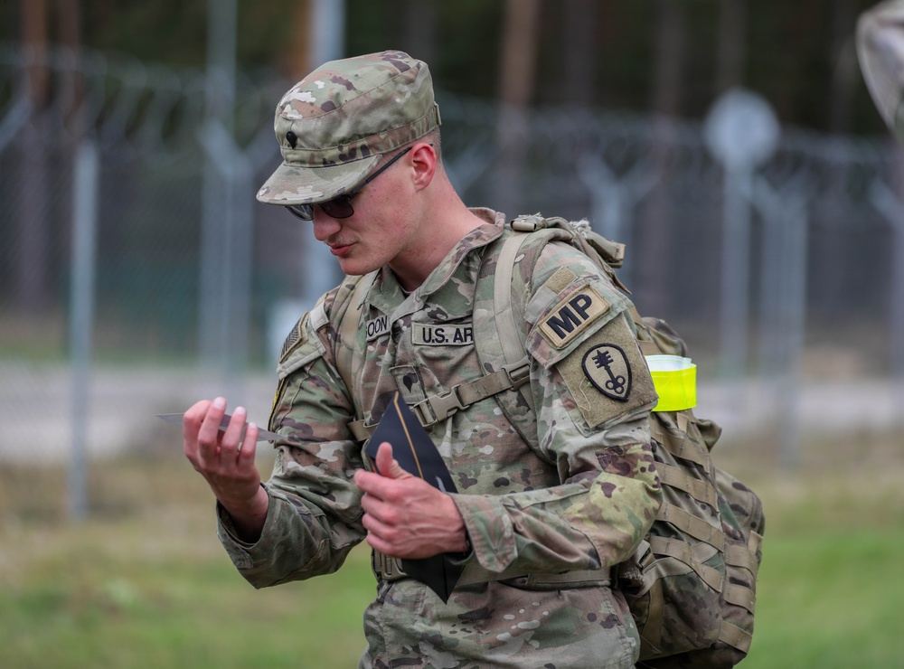 A U.S. Army Military Police (MP) soldier in OCP (Operational Camouflage Pattern) uniform is examining a small object or card while standing outdoors. The soldier is wearing full gear including a rucksack and has an 'MP' patch and a unit insignia patch visible on the left shoulder. A yellow high-visibility band is attached to the backpack strap, suggesting a training or exercise environment.