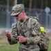 A U.S. Army Military Police (MP) soldier in OCP (Operational Camouflage Pattern) uniform is examining a small object or card while standing outdoors. The soldier is wearing full gear including a rucksack and has an 'MP' patch and a unit insignia patch visible on the left shoulder. A yellow high-visibility band is attached to the backpack strap, suggesting a training or exercise environment.