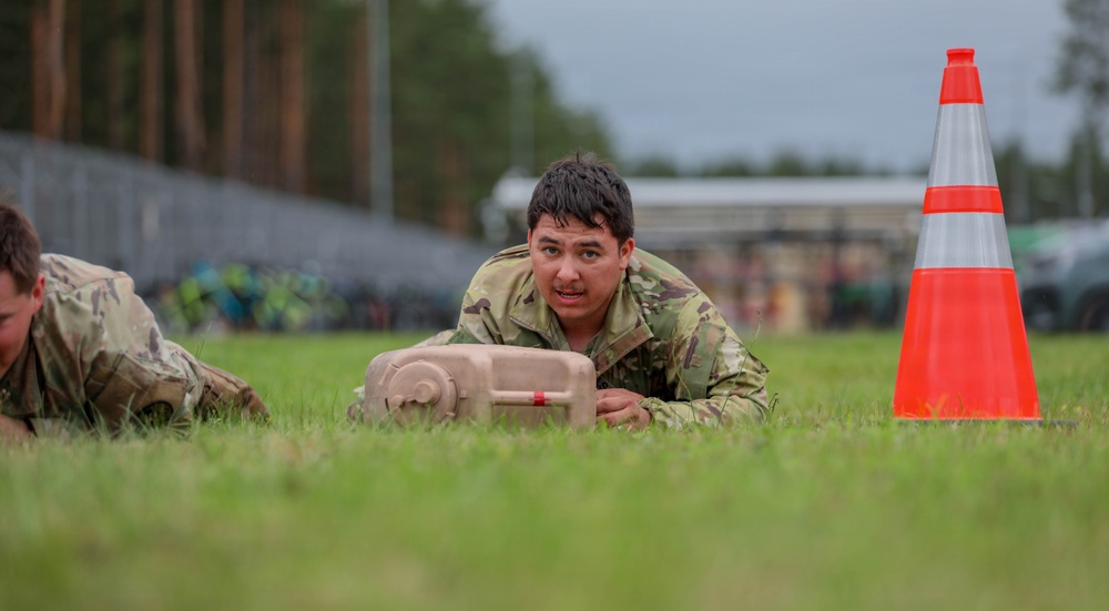 A soldier in camouflage uniform is low-crawling across a grass field during what appears to be a military training exercise or competition. The soldier is pushing or carrying a tan-colored training aid object while crawling on their stomach. A second soldier is partially visible on the left, and an orange traffic cone marks a boundary or course marker nearby.
