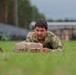 A soldier in camouflage uniform is low-crawling across a grass field during what appears to be a military training exercise or competition. The soldier is pushing or carrying a tan-colored training aid object while crawling on their stomach. A second soldier is partially visible on the left, and an orange traffic cone marks a boundary or course marker nearby.