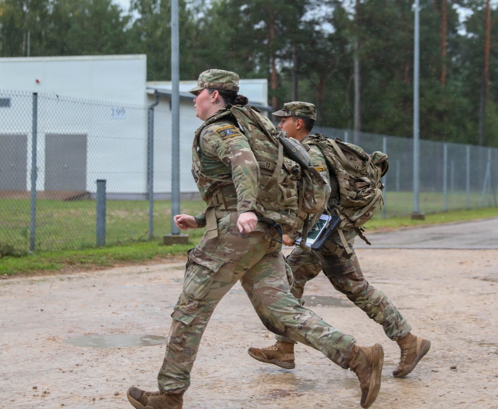 Two soldiers in US Army OCP (Operational Camouflage Pattern) uniforms are running on a paved surface at what appears to be a military installation or training base. Both are carrying large rucksacks and wearing patrol caps, suggesting a ruck march or physical training exercise. The setting appears to be in a northern European environment based on the vegetation and infrastructure visible in the background.