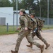 Two soldiers in US Army OCP (Operational Camouflage Pattern) uniforms are running on a paved surface at what appears to be a military installation or training base. Both are carrying large rucksacks and wearing patrol caps, suggesting a ruck march or physical training exercise. The setting appears to be in a northern European environment based on the vegetation and infrastructure visible in the background.