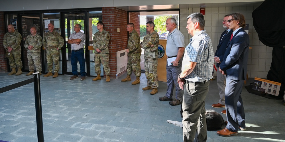 A group of approximately ten individuals are standing in a line inside what appears to be a military facility lobby or entrance area. The group consists of a mix of uniformed military personnel in camouflage (OCP/MultiCam) uniforms and civilians in business or casual attire. They appear to be attending a formal briefing, ceremony, or official visit.
