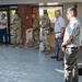 A group of approximately ten individuals are standing in a line inside what appears to be a military facility lobby or entrance area. The group consists of a mix of uniformed military personnel in camouflage (OCP/MultiCam) uniforms and civilians in business or casual attire. They appear to be attending a formal briefing, ceremony, or official visit.