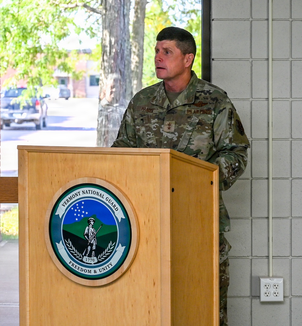 A U.S. Air Force officer, wearing OCP (Operational Camouflage Pattern) uniform with a two-star Major General rank insignia, is speaking at a podium bearing the Vermont National Guard seal. The seal displays the motto 'Freedom &amp; Unity' and the year 1770. The setting appears to be an indoor/covered outdoor venue with natural light visible through windows in the background.