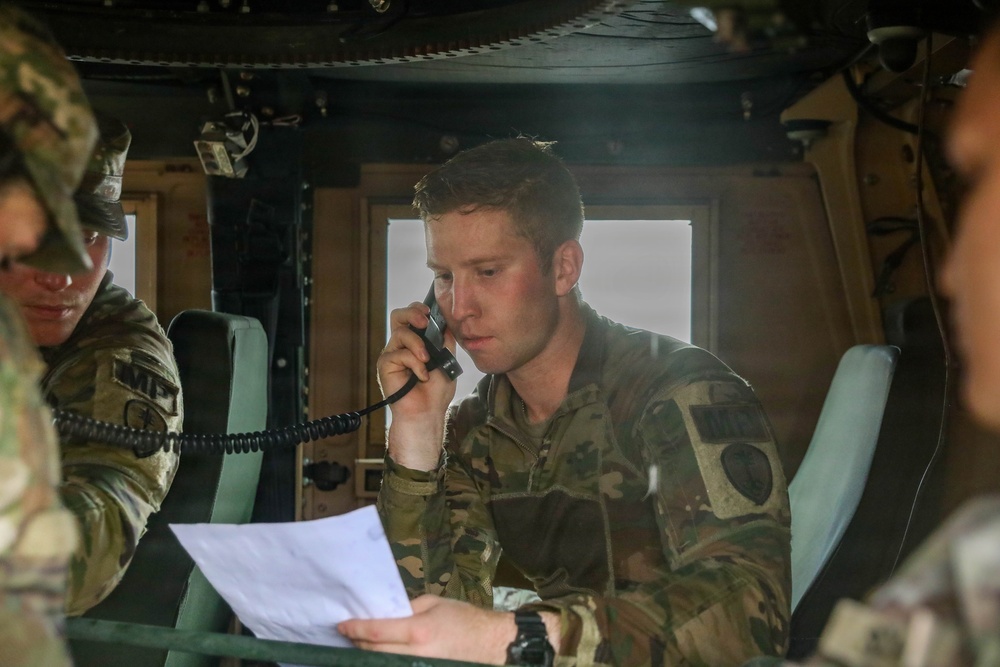 Military personnel, identifiable by 'MP' (Military Police) patches on their uniforms, are seated inside what appears to be a military vehicle such as a HMMWV (Humvee). The central figure is using a corded radio handset while referencing a document, suggesting active communication during an operation or exercise. At least two other soldiers are visible in the frame.