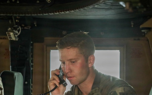Military personnel, identifiable by 'MP' (Military Police) patches on their uniforms, are seated inside what appears to be a military vehicle such as a HMMWV (Humvee). The central figure is using a corded radio handset while referencing a document, suggesting active communication during an operation or exercise. At least two other soldiers are visible in the frame.