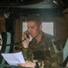 Military personnel, identifiable by 'MP' (Military Police) patches on their uniforms, are seated inside what appears to be a military vehicle such as a HMMWV (Humvee). The central figure is using a corded radio handset while referencing a document, suggesting active communication during an operation or exercise. At least two other soldiers are visible in the frame.
