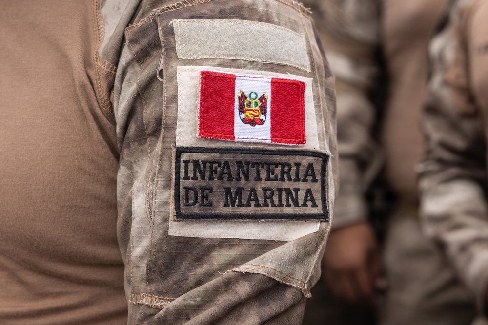 Close-up photograph of a military uniform sleeve displaying two patches on a desert camouflage (MultiCam/arid pattern) uniform. The patches identify the wearer as a member of the Peruvian Marine Corps (Infantería de Marina). A Peruvian national flag patch with the coat of arms is prominently displayed above the unit identification patch.