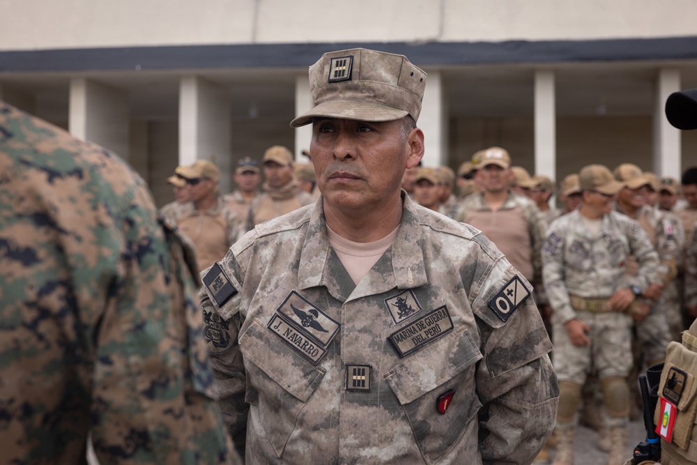 A senior officer of the Marina de Guerra del Perú (Peruvian Navy) identified by name tape as 'J. Navarro' stands at attention during what appears to be a military formation or ceremony. He wears a camouflage uniform with rank insignia on his cap and chest, along with a naval warfare qualification badge. Behind him, rows of uniformed military personnel are visible, along with what appears to be a U.S. Marine in woodland MARPAT camouflage on the left.
