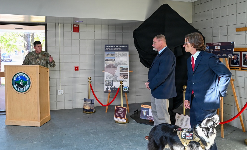 A formal military ceremony is taking place in what appears to be a building lobby or hallway of the Vermont National Guard. A uniformed soldier is speaking at a podium while two civilians in suits stand near a partially unveiled display covered in black cloth. The event appears to be a dedication or unveiling ceremony related to 'Task Force Saber' and Vermont National Guard history.