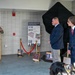 A formal military ceremony is taking place in what appears to be a building lobby or hallway of the Vermont National Guard. A uniformed soldier is speaking at a podium while two civilians in suits stand near a partially unveiled display covered in black cloth. The event appears to be a dedication or unveiling ceremony related to 'Task Force Saber' and Vermont National Guard history.