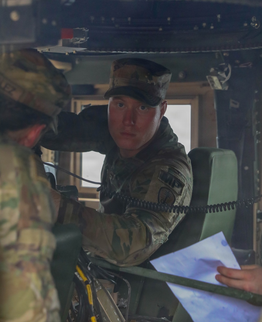 Two soldiers in U.S. Army Operational Camouflage Pattern (OCP) uniforms are inside what appears to be a military vehicle, likely a HMMWV (Humvee). One soldier is seated and looking directly at the camera while holding a coiled communication cord, while another soldier (partially visible) holds printed documents. They appear to be conducting a briefing or reviewing operational materials.