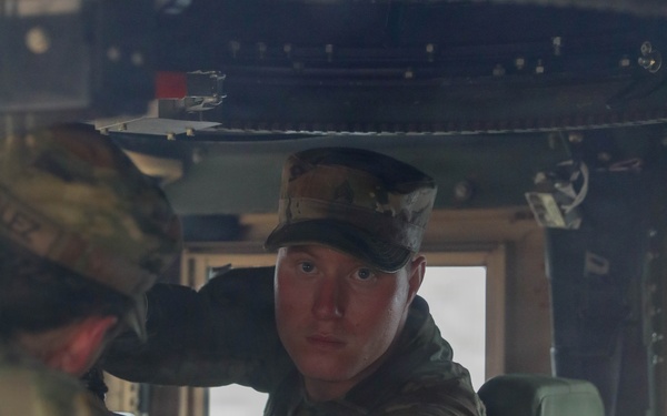 Two soldiers in U.S. Army Operational Camouflage Pattern (OCP) uniforms are inside what appears to be a military vehicle, likely a HMMWV (Humvee). One soldier is seated and looking directly at the camera while holding a coiled communication cord, while another soldier (partially visible) holds printed documents. They appear to be conducting a briefing or reviewing operational materials.
