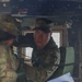 Two soldiers in U.S. Army Operational Camouflage Pattern (OCP) uniforms are inside what appears to be a military vehicle, likely a HMMWV (Humvee). One soldier is seated and looking directly at the camera while holding a coiled communication cord, while another soldier (partially visible) holds printed documents. They appear to be conducting a briefing or reviewing operational materials.