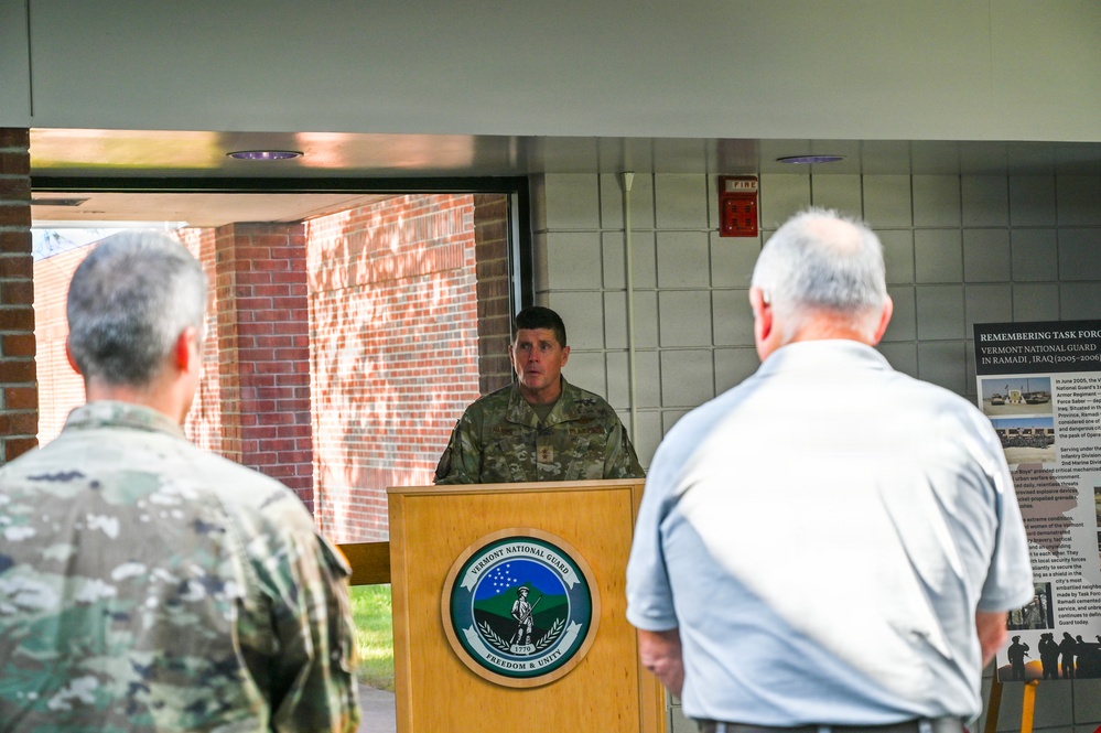 A military officer in Army Combat Uniform (ACU) is speaking at a podium bearing the Vermont National Guard seal with the motto 'Freedom &amp; Unity'. The event appears to be a memorial or commemorative ceremony, as evidenced by a display panel visible in the background titled 'Remembering Task Force' referencing Vermont National Guard operations in Ramadi, Iraq (2005-2006). Two attendees are visible from behind in the foreground, one in military uniform and one in civilian clothing.