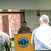 A military officer in Army Combat Uniform (ACU) is speaking at a podium bearing the Vermont National Guard seal with the motto 'Freedom &amp; Unity'. The event appears to be a memorial or commemorative ceremony, as evidenced by a display panel visible in the background titled 'Remembering Task Force' referencing Vermont National Guard operations in Ramadi, Iraq (2005-2006). Two attendees are visible from behind in the foreground, one in military uniform and one in civilian clothing.
