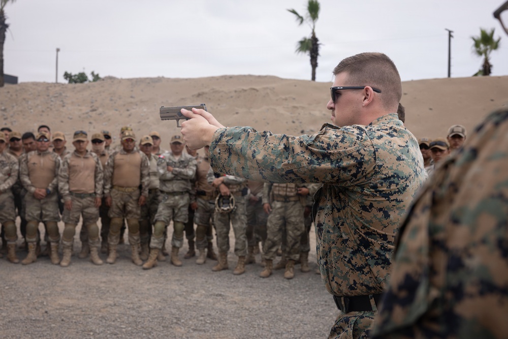 A military instructor in USMC MARPAT digital camouflage demonstrates pistol handling technique to a large group of assembled soldiers at an outdoor shooting range. The instructor is in a two-handed shooting stance, aiming a semi-automatic pistol while a formation of troops in mixed camouflage uniforms observes. The setting appears to be a training facility with sand berms, palm trees, and an overcast sky.