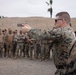 A military instructor in USMC MARPAT digital camouflage demonstrates pistol handling technique to a large group of assembled soldiers at an outdoor shooting range. The instructor is in a two-handed shooting stance, aiming a semi-automatic pistol while a formation of troops in mixed camouflage uniforms observes. The setting appears to be a training facility with sand berms, palm trees, and an overcast sky.