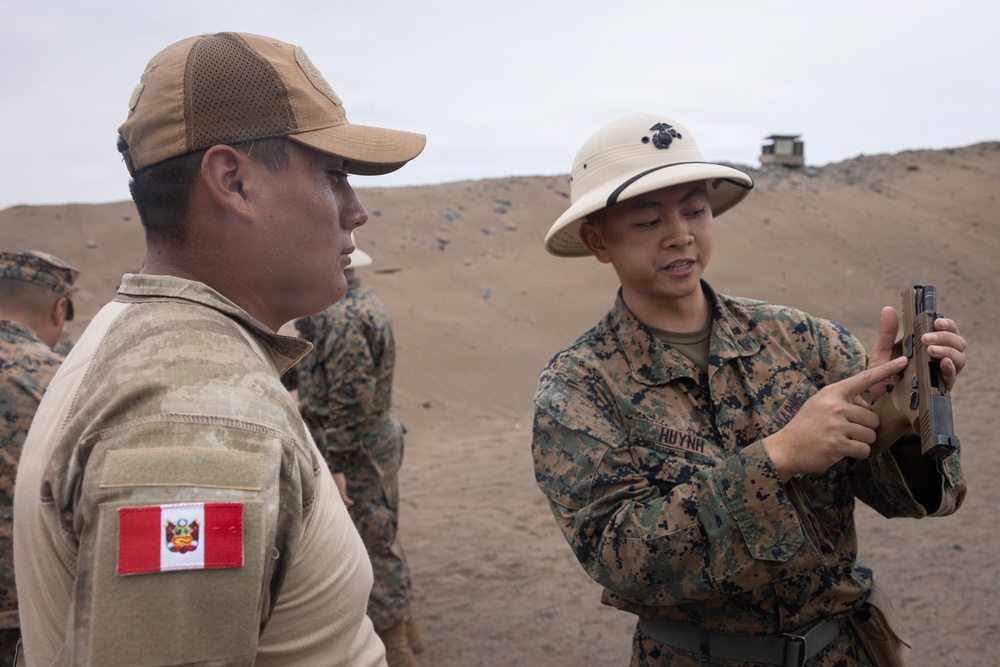 A U.S. Marine (identified by name tape 'HUYNH' and MARPAT camouflage uniform) is demonstrating or explaining a pistol to a Peruvian military personnel (identified by the Peruvian flag patch on the left shoulder). The interaction appears to take place on a sandy outdoor firing range or training area. This appears to be a joint military training or exchange exercise between U.S. Marines and Peruvian armed forces.
